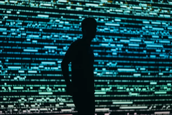 Silhouette of a man in front of a large screen with lines of code on it.